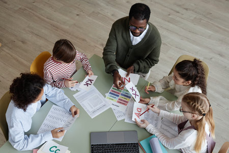 From above view shot of young Black man sitting at table with kids in classroom teaching English language, copy spaceの写真素材
