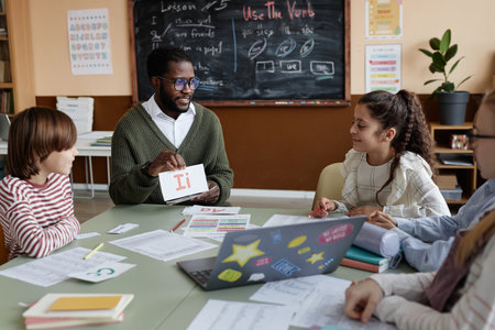 Young African American teacher of English sitting at table in classroom showing alphabet cards to kids during lessonの写真素材