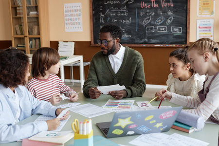 Young African American male teacher of English sitting at table in classroom working with...