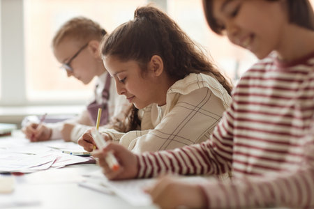 Selective focus shot of Middle Eastern girl sitting at table with her classmates making notes during lessonの写真素材