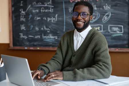 Medium portrait of young African American male teacher of English wearing eyeglasses sitting at desk in classroom smiling at camera while using laptopの写真素材