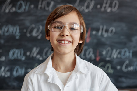 Closeup portrait of cheerful Caucasian schoolboy wearing lab coat and goggles standing against blackboard in classroom smiling at cameraの写真素材
