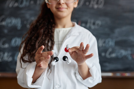 Medium closeup of schoolgirl wearing lab coat standing against blackboard holding chemical structure model in handsの写真素材