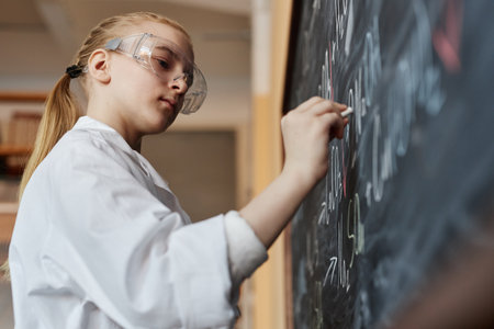 Low angle view of serious girl wearing goggles and lab coat writing formula on blackboard in Chemistry class, copy spaceの写真素材