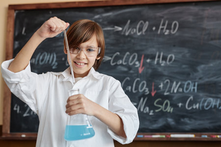 Medium portrait of cheerful Caucasian boy wearing lab coat and goggles holding flask with blue liquid standing against blackboard in Chemistry classの写真素材