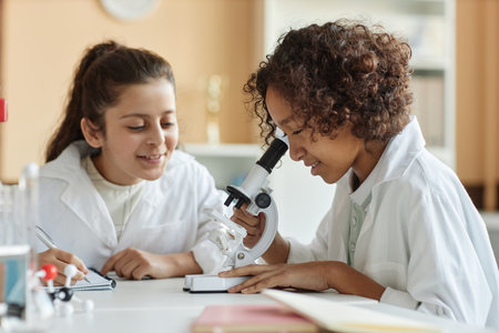 African American boy wearing lab coat sitting in classroom looking through microscope while Middle Eastern girl making notes in notebookの写真素材