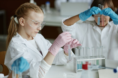 Selective focus shot of Caucasian girl sitting at table in classroom mixing liquids in test tube...