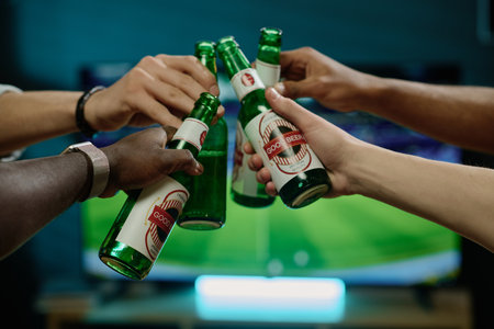 Closeup shot of hands of unrecognizable men clinking bottles with beer while watching soccer game on...