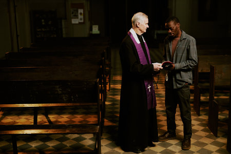Long shot of ethnically diverse senior Catholic priest and young Black man standing indoors discussing religious text, copy spaceの写真素材
