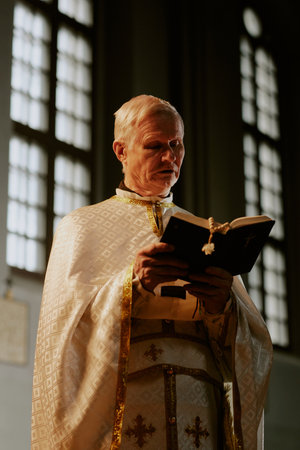 Vertical low angle medium shot of senior Catholic priest wearing liturgical vestment reading holy Bible book, copy spaceの写真素材
