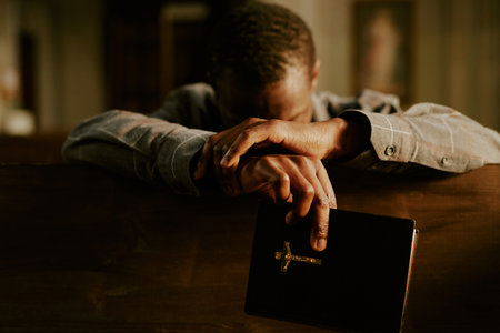 Medium closeup of unrecognizable Black man sitting in Catholic church with Bible in hand praying to...