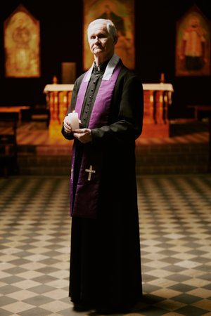 Vertical long shot of senior Catholic priest wearing cassock with stole holding candle in hands standing in church looking at cameraの写真素材