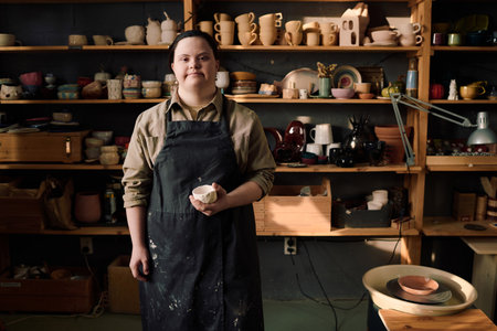 Medium long shot of young Caucasian female artisan with Down syndrome standing in pottery workshop...