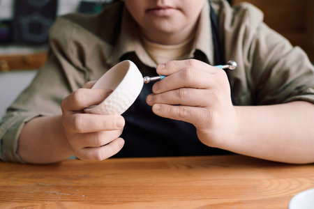 Closeup of hands of woman with Down syndrome using modeling ball tool while working on ceramic dishの写真素材
