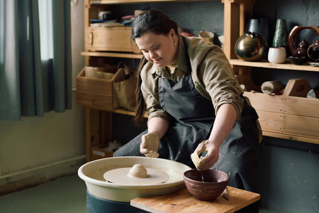 Medium long shot of young woman with Down syndrome wearing apron sitting at potters wheel taking...