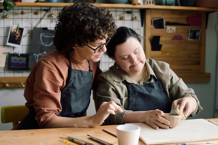 Medium shot of mature Caucasian woman teaching student with Down syndrome how to make ceramic bowl...