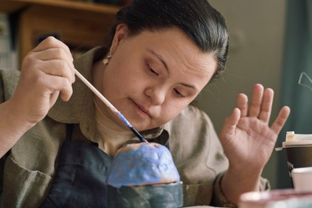 Creative young woman with Down syndrome sitting at table in workshop coloring handmade vessel with...