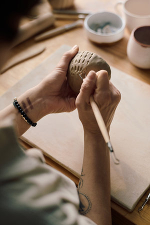 Vertical high angle shot of hands of unrecognizable artisan using clay carving tool to decorate...