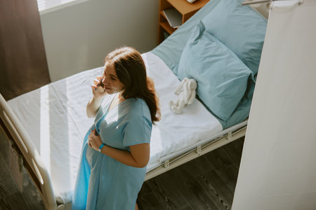 High angle photo of young pregnant woman walking around hospital ward and holding her belly while she speaking on phoneの写真素材