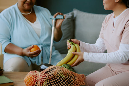 Young nurse treating senior African American female patient to bananas while patient sitting on couch holding ripe orange and smiling sincerelyの写真素材