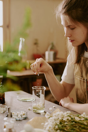 Young female perfumer strewing petals of dried crushed daisy into transparent aromatic liquidの写真素材