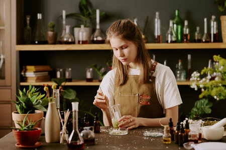 Female young perfumer mixing transparent liquid with dried wildflowers carefully while she in home perfume lab making new types of perfume and cologneの写真素材