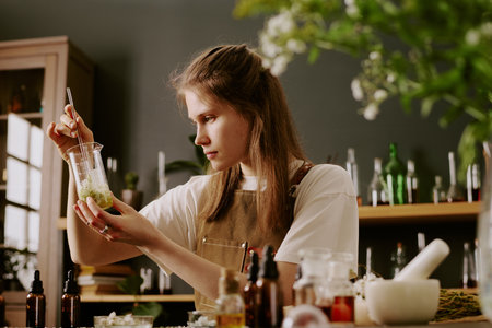 Young female perfumer sitting at work desk with perfume craft items and admiring at golden liquid with wildflowers petals while she mixing itの写真素材