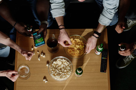 From above view of unrecognizable friends eating snacks, drinking beer and betting in online app while watching sports eventの写真素材