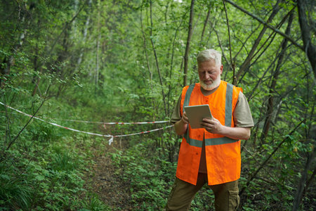 Senior Caucasian ecologist wearing orange vest working on digital tablet in forest, copy space