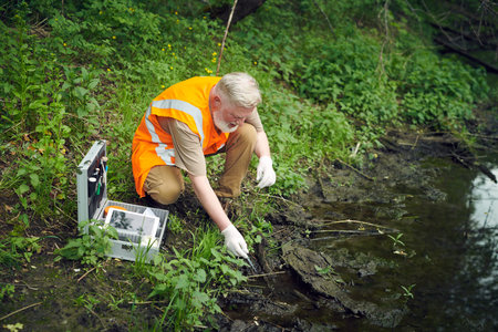 High angle view of senior scientist wearing orange safety vest taking water sample from river with...