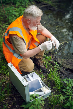 Vertical high angle view of senior male ecologist wearing safety vest with gloves taking river water...
