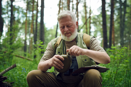 Medium shot of mature Caucasian traveler opening water flask while having rest during hiking in...