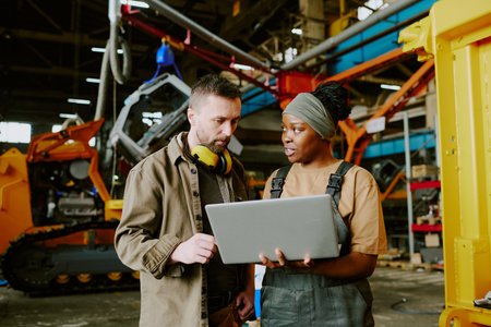 Medium shot of biracial male and female factory workers looking at laptop screen and discussing new...