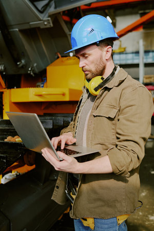 Vertical medium shot of Caucasian technician wearing hardhat typing on laptop at work in machinery...