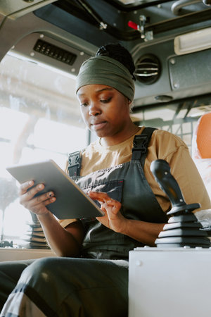 Vertical medium shot of young Black female factory worker sitting inside tractor using digital...