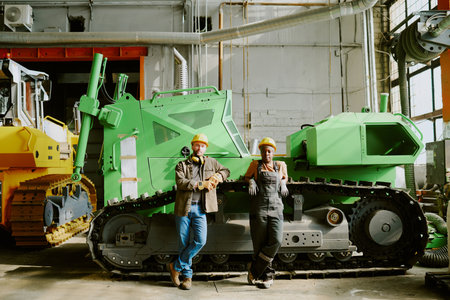 Wide shot of Black woman and Caucasian man in workwear standing relaxed against green bulldozer...