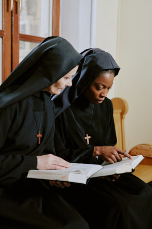 Vertical medium shot of two biracial nuns sitting on wooden bench in Catholic church and discussing...
