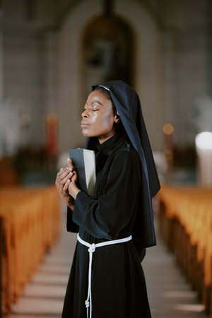 Vertical medium shot of pious young African American nun standing with eyes closed in church nave with religious book in handsの写真素材