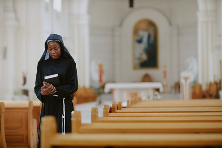 Medium long shot of young African American nun holding Bible book walking along nave in Catholic church, copy spaceの写真素材