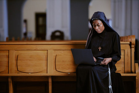 Medium shot of young African American nun sitting on wooden pew typing on laptop, copy spaceの写真素材