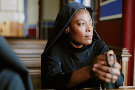 Young African American Catholic nun holding rosary beads sitting on pew and looking away, medium closeup shotの写真素材