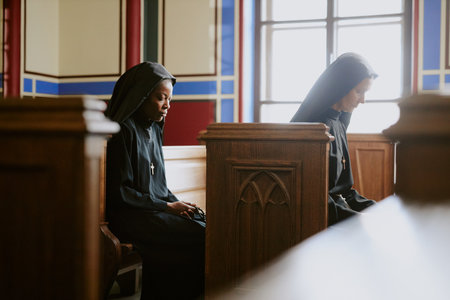 Side view shot of two diverse nuns sitting on wooden benches in Catholic cathedral or church praying...