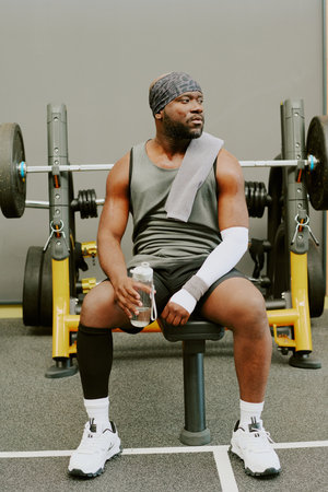 African American man sitting on leather bench and drinking pure water after workout
