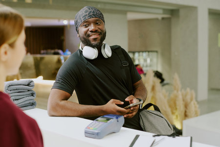 Young African American gym resident talking to female receptionist in huge fitness club lobby