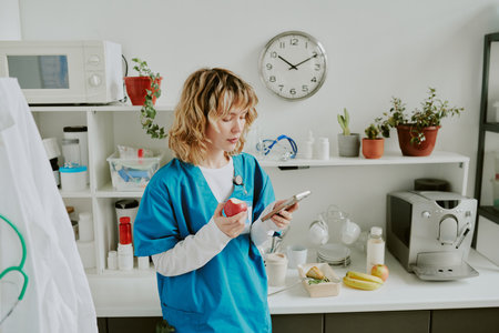 Young pretty Caucasian nurse eating red apple while communicating via smartphone during her break at hospitalの写真素材