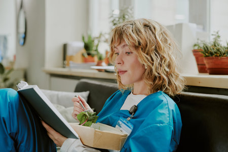 Profile shot of young Caucasian nurse enjoying novels lines while eating salad in on call room...