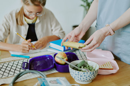 Unrecognizable female hands preparing lunch box and putting healthy burrito and salad in it while daughter drawingの写真素材