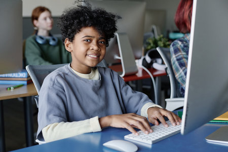Medium shot of cheerful African American boy sitting at desk in modern classroom smiling at camera...