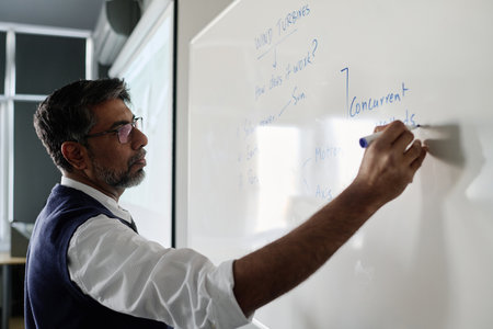 Medium closeup of mature biracial teacher wearing eyeglasses writing on board during lesson, copy...