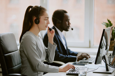 Young Caucasian operator speaking with client via wired headphone while his African American coworker searching information in program in blurred backgroundの写真素材